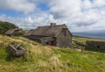 Traditional Irish stone cottage in the countryside with thatched roof and white-painted walls under a summer sky.