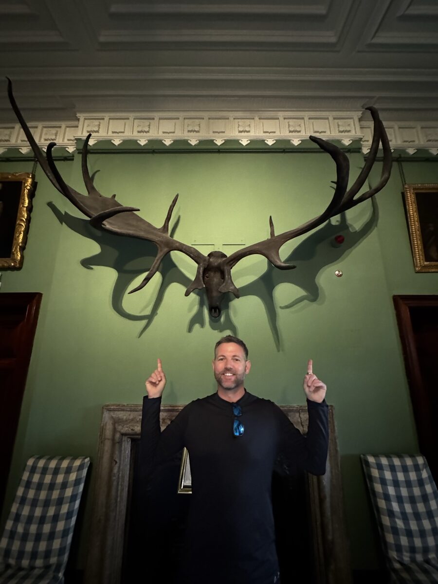 Man posing beneath massive ancient Irish elk antlers mounted on green wall in Ireland castle