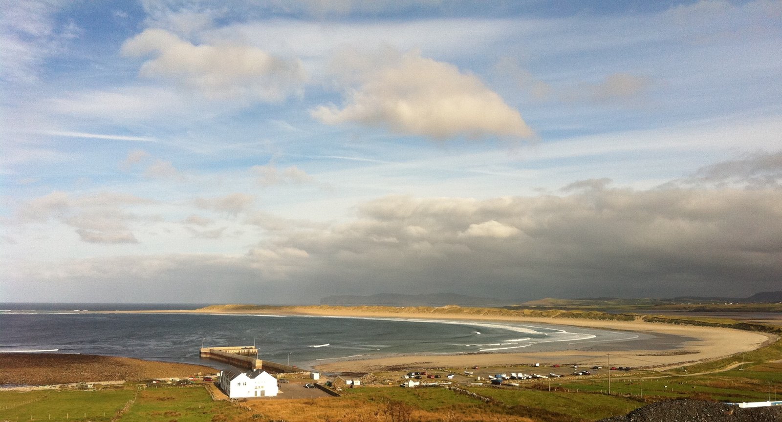 Wide curved sandy bay with green fields and a small white building on the wild Donegal coast.