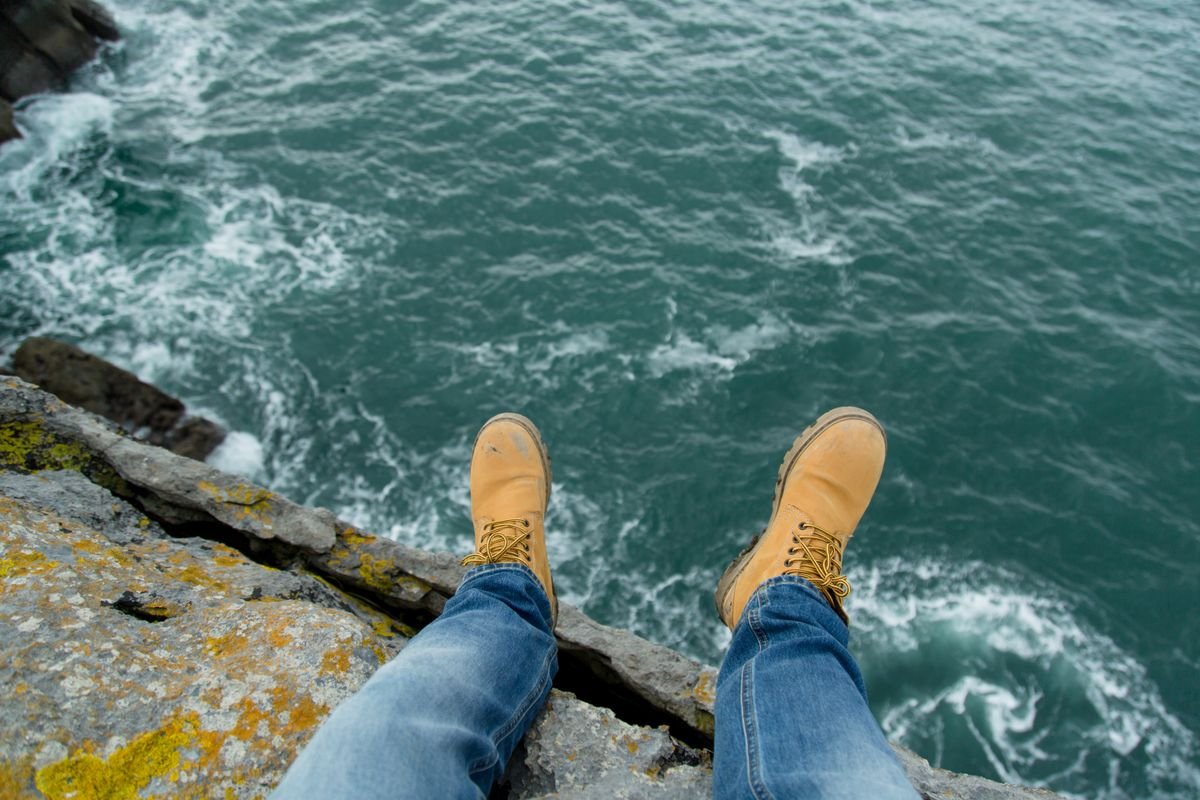 A walker's feet at the unguarded edge of a sea cliff with the Atlantic churning far below