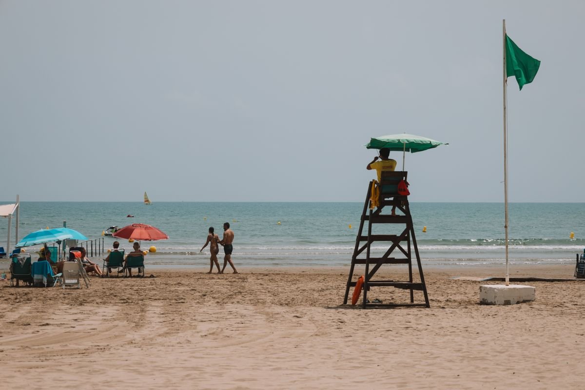 A lifeguard tower and a green safety flag on a sandy beach with people swimming