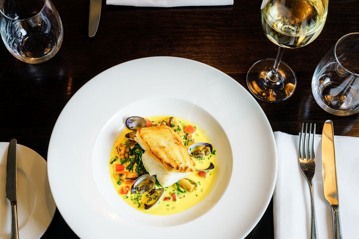 A plated fish dish with mussels and a glass of white wine on a restaurant table