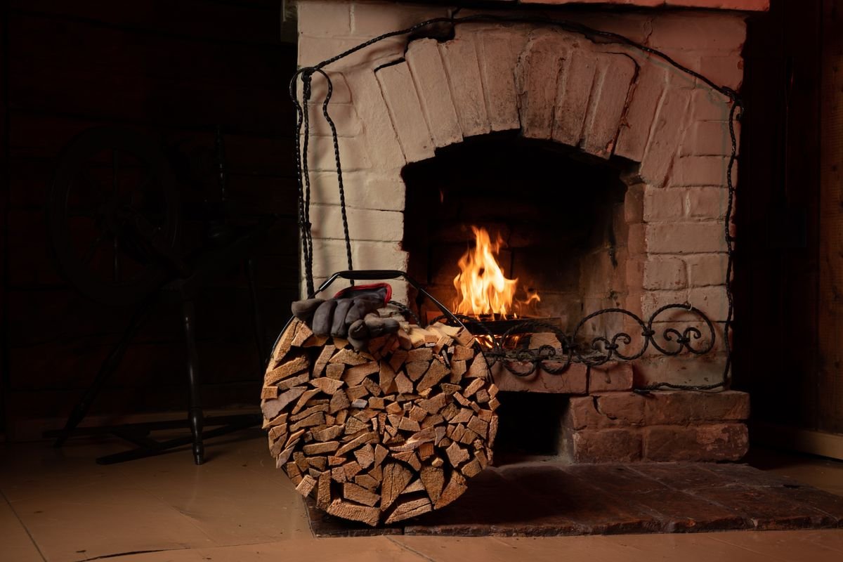 A rustic brick fireplace with a lit fire and a basket of chopped logs