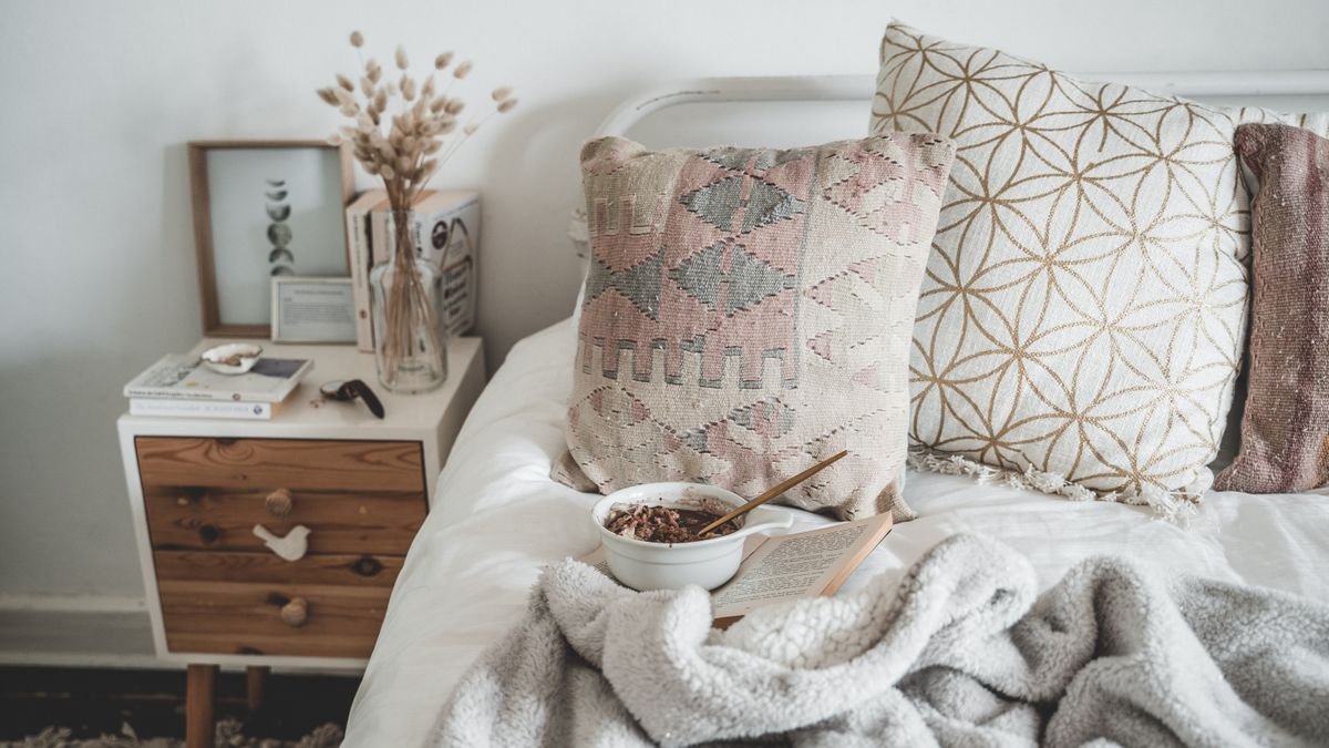 A cosy bed with a soft throw blanket, knit cushions and books on a bedside table
