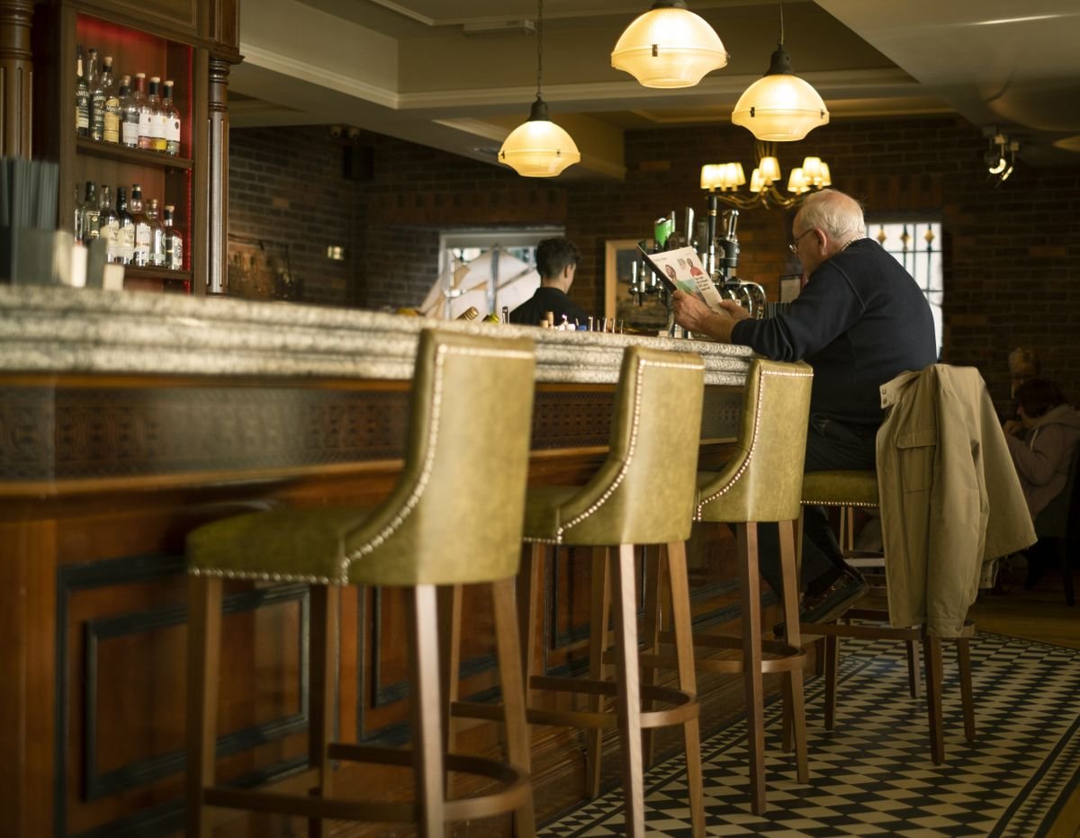 Stools lined up along a warm wooden pub counter with a man reading at the bar