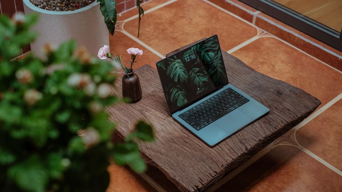 A laptop on a wooden table beside greenery in a bright room