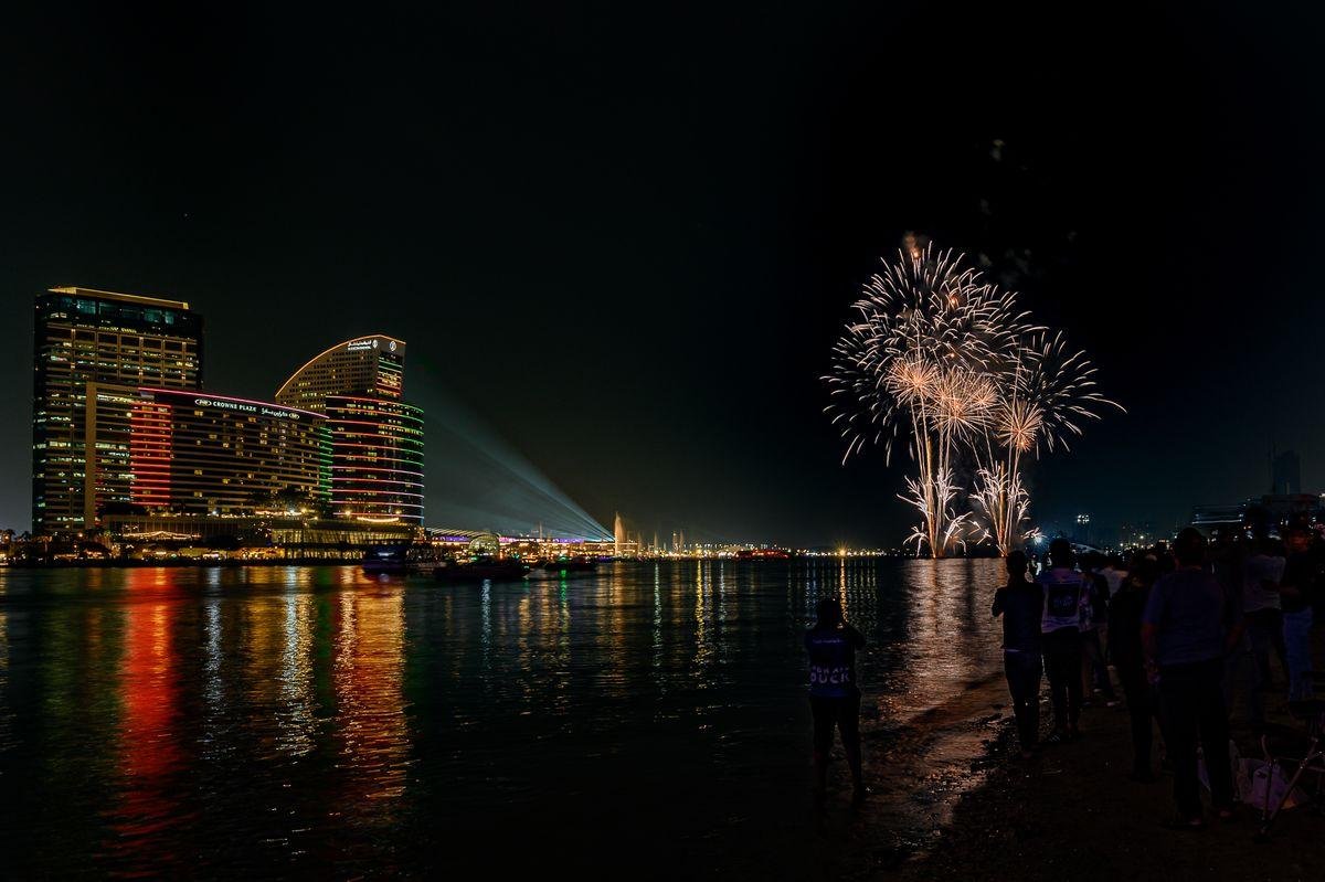 Fireworks bursting over a river with a crowd watching from the bank