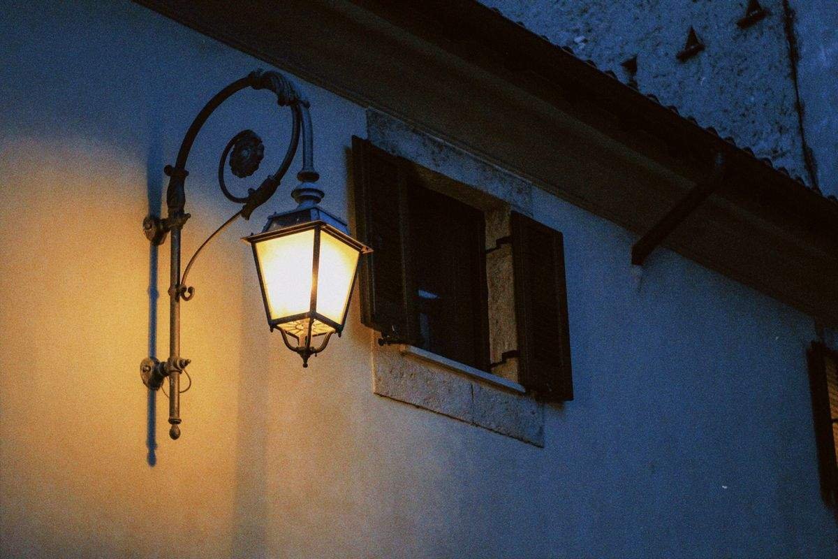 An old wall mounted lantern glowing against a stone building at dusk