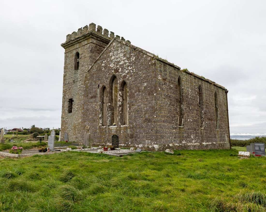 A ruined stone church with a battlemented tower beside a coastal graveyard