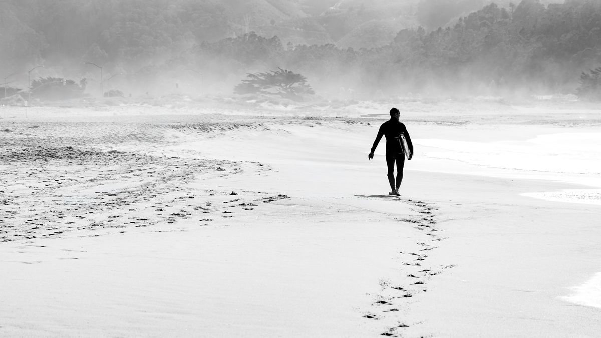 A lone surfer in a wetsuit walking a misty empty beach carrying a board