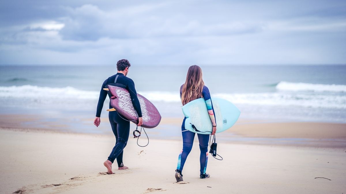 Two surfers in wetsuits walking toward a grey ocean carrying boards