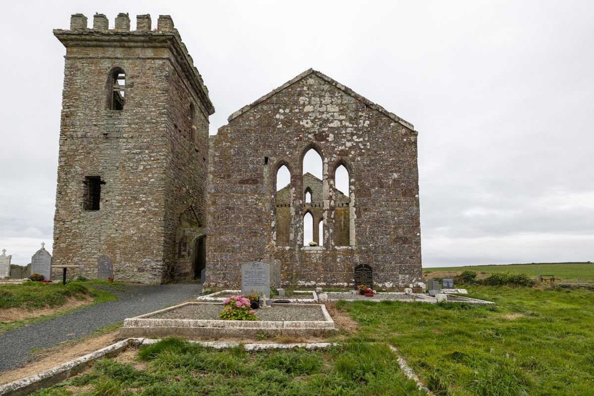 A roofless stone friary church with a battlemented tower and Gothic windows in a graveyard