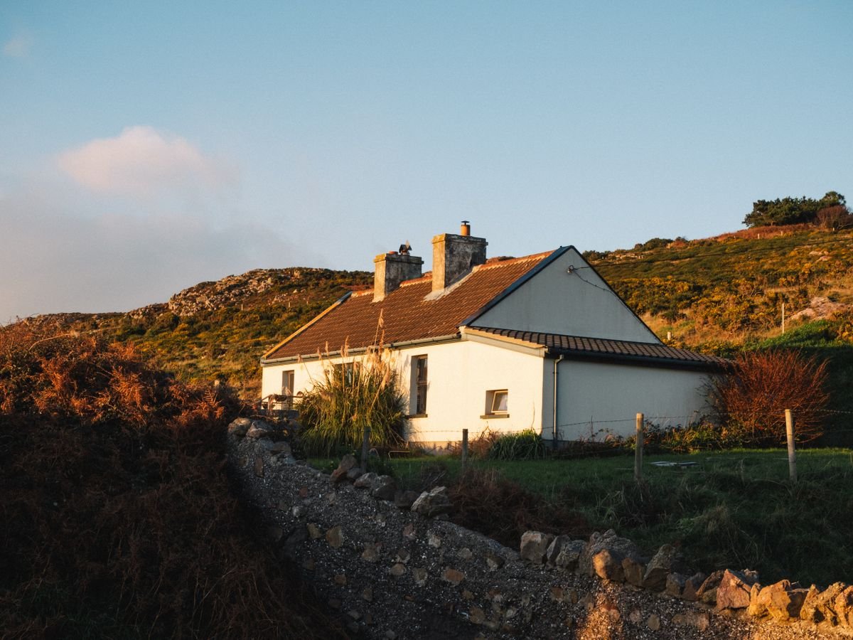 A whitewashed Irish cottage with a stone wall on a grassy Donegal hillside at golden hour