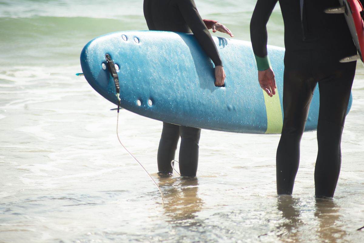Two surfers in black wetsuits holding a foam board in shallow water