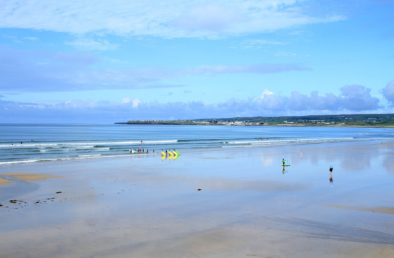 The long flat beach at Lahinch in County Clare at low tide with surf schools carrying boards and the town in the distance