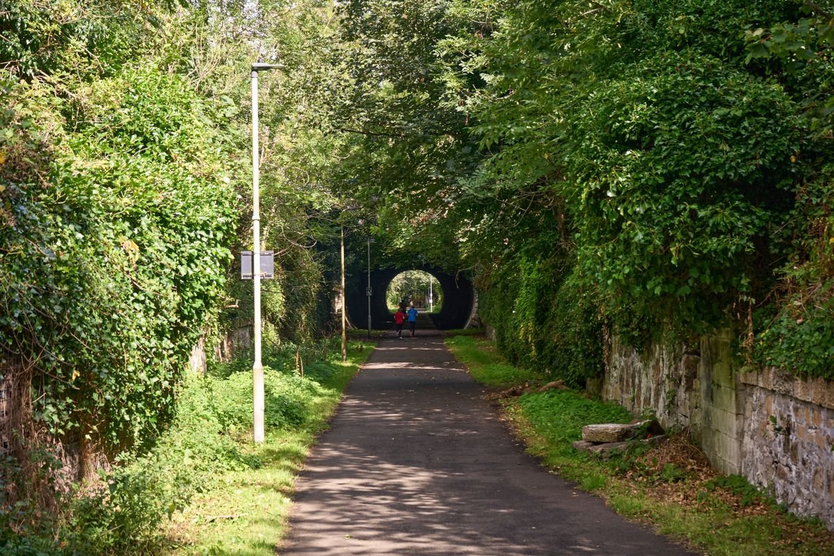 A paved greenway path leading to an old railway tunnel through wooded cuttings