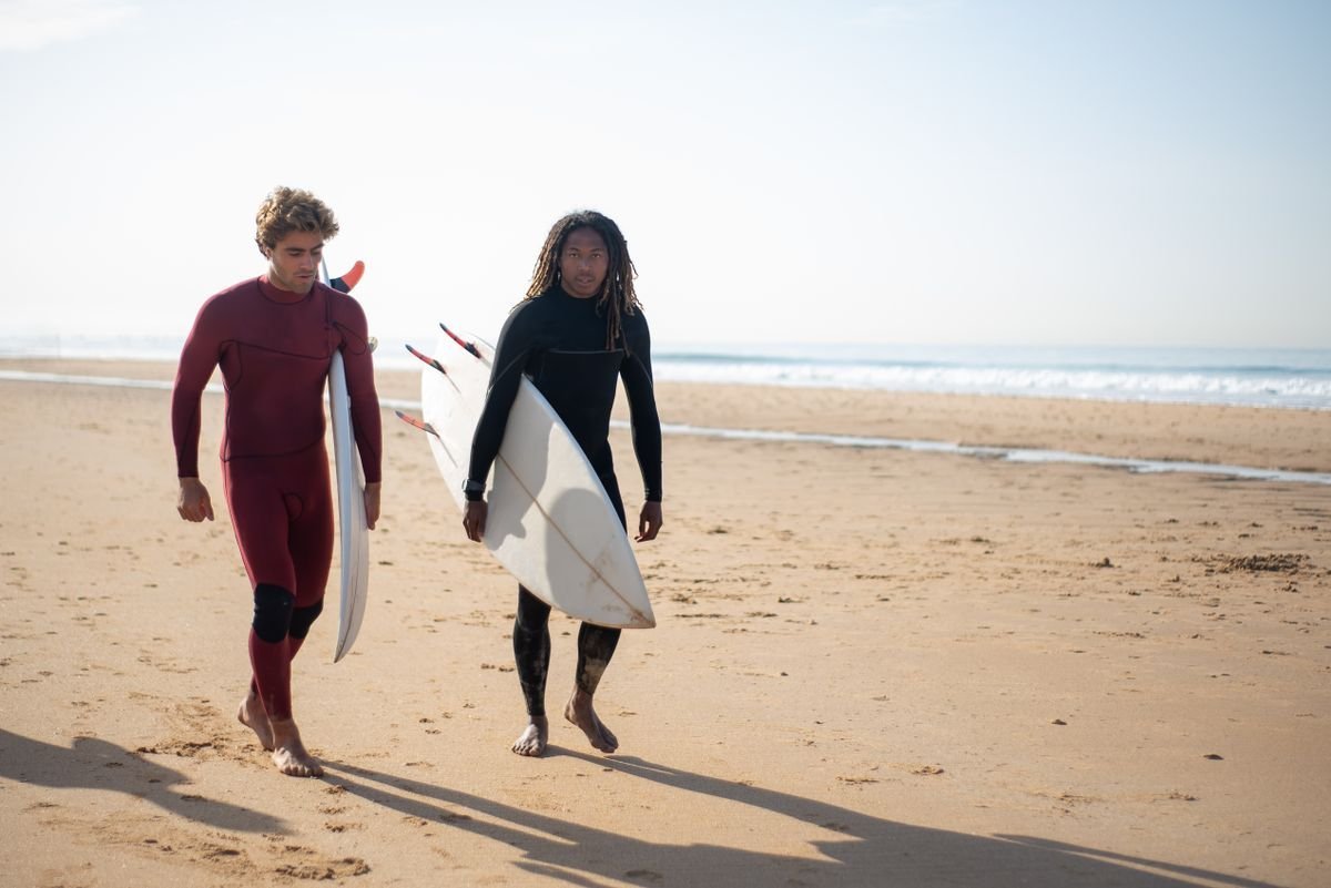 Two surfers in wetsuits carrying surfboards along a sandy beach