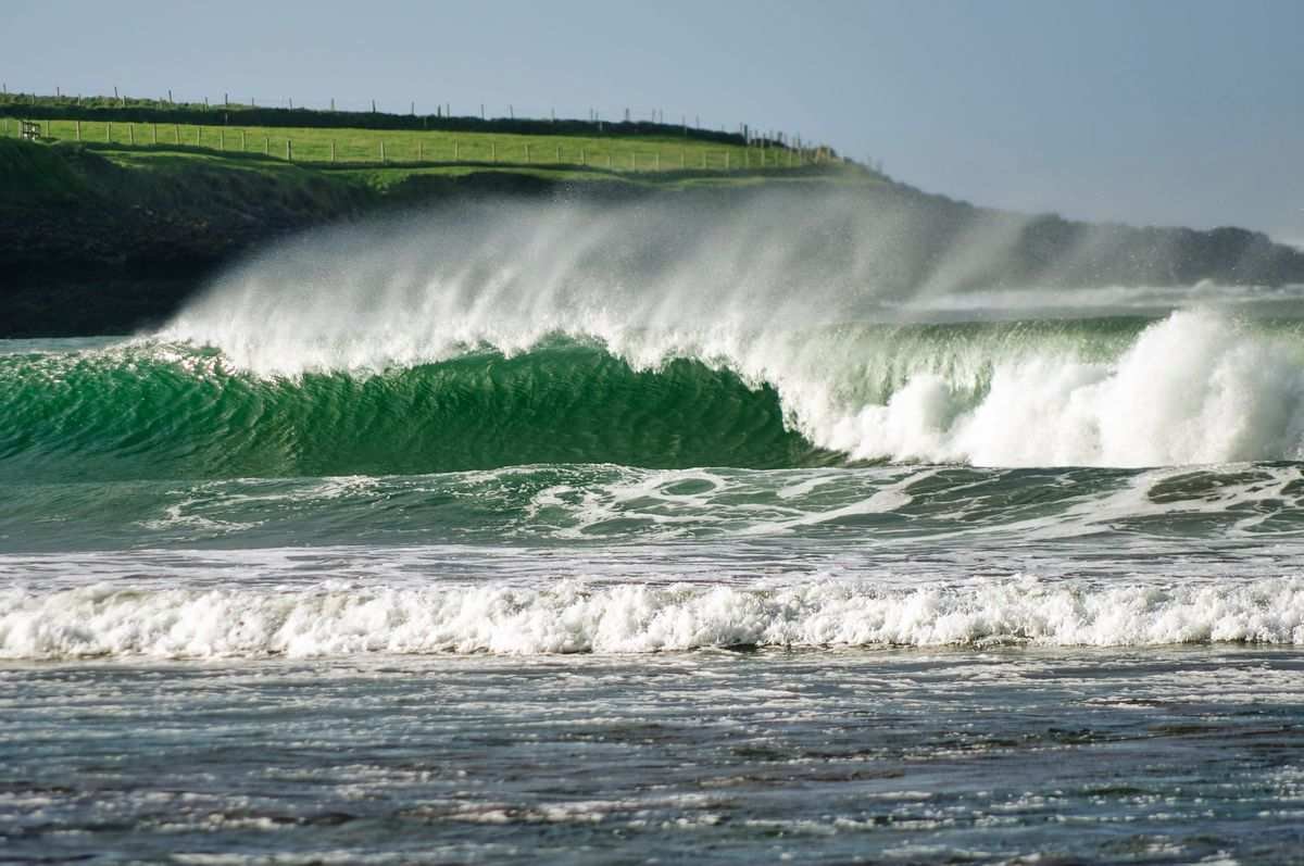 A large green Atlantic wave breaking with a grassy Irish headland behind