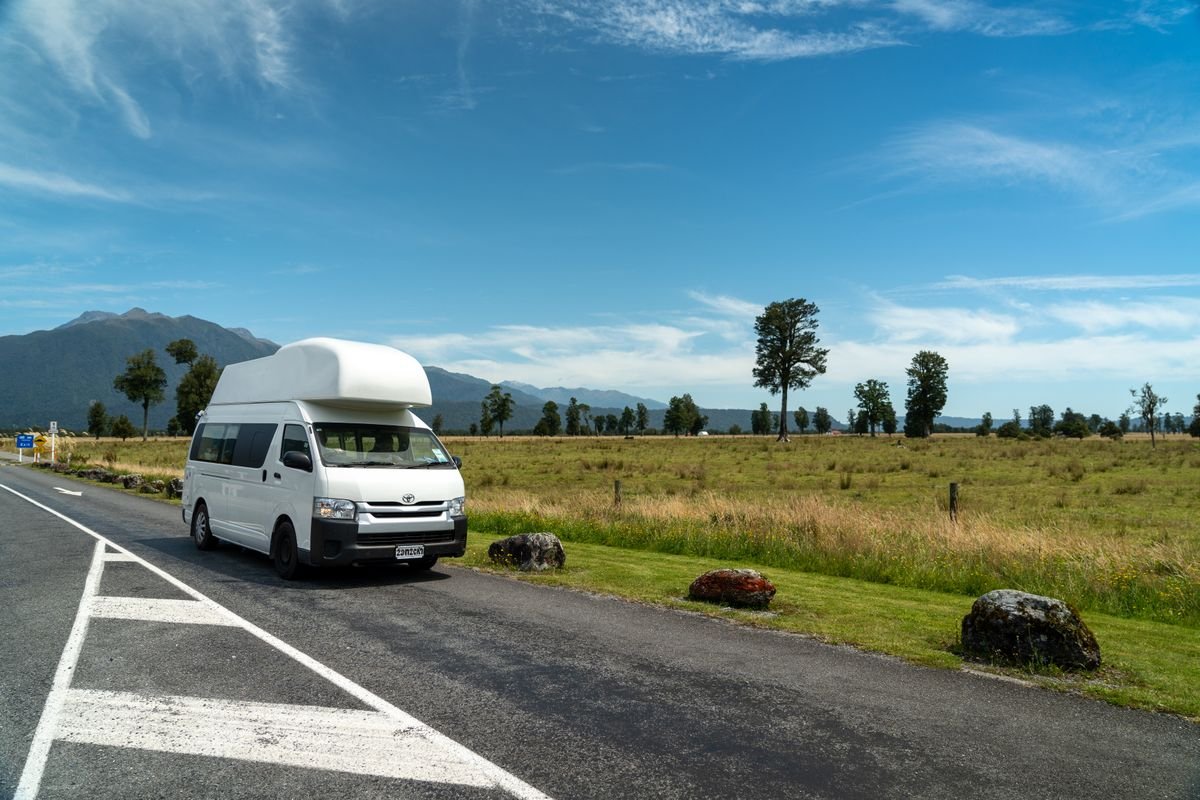 A white campervan parked on a quiet rural road with grassland and mountains behind.