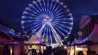 A lit Ferris wheel rising over a crowded Christmas market at dusk