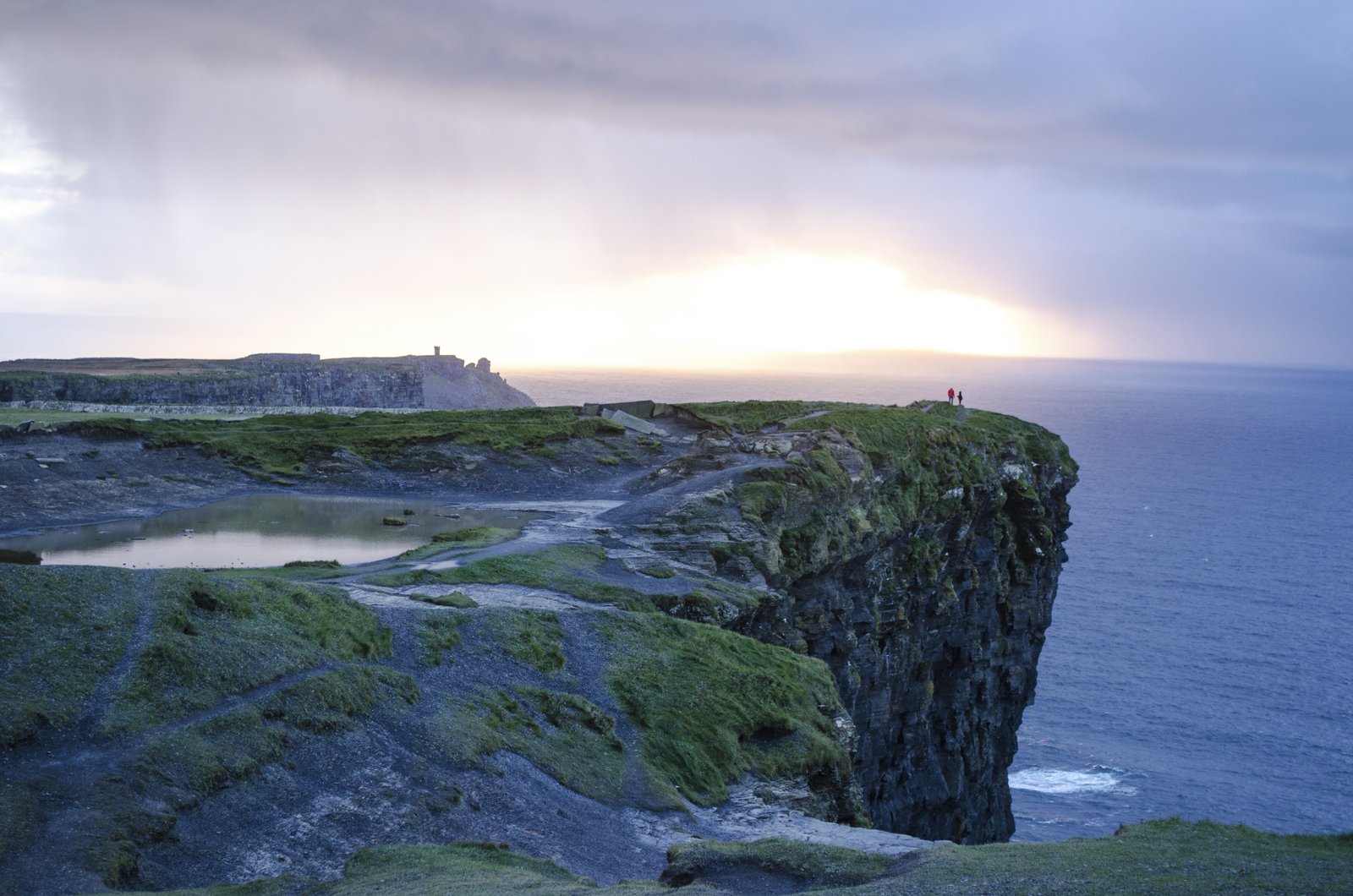 A rain shower crossing the Cliffs of Moher at sunset with two people on the clifftop path